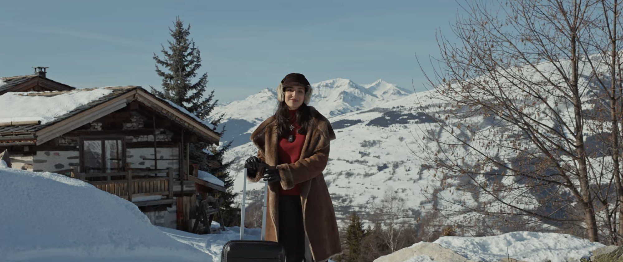 Voyageuse avec sa valise devant un chalet enneigé dans les Alpes
